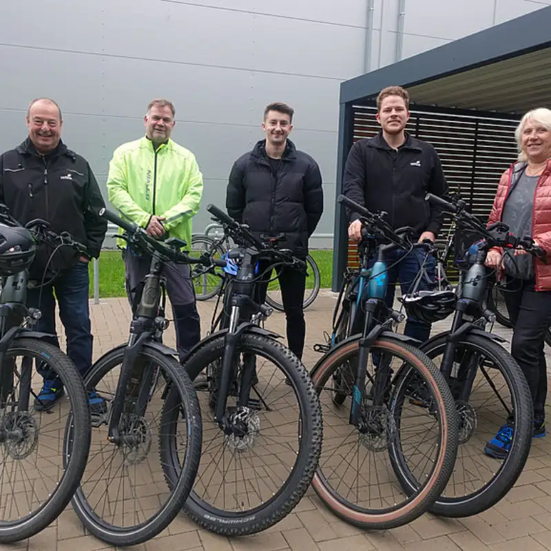 Fünf Hettich Mitarbeitende stehen, bei trübem Wetter, stehen zusammen mit ihren Fahrrädern vor einem Fahrradstand Fünf Hettich Mitarbeitende stehen, bei trübem Wetter, stehen zusammen mit ihren Fahrrädern vor einem Fahrradstand