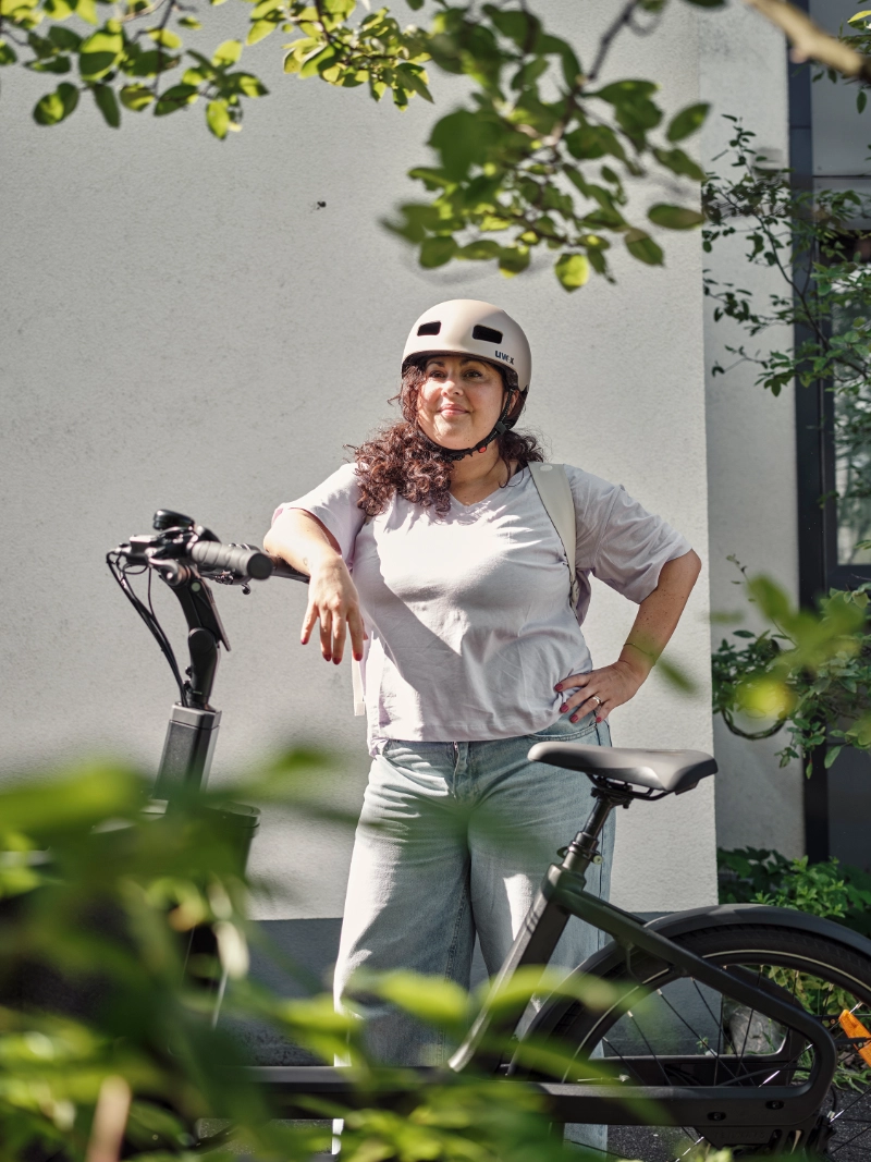 Frau mit Locken lehnt am JobRad, im Vordergrund Pflanzen Frau mit Locken lehnt am JobRad, im Vordergrund Pflanzen